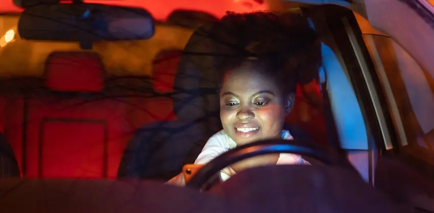 A young woman smiles at the glowing screen of her phone while seated behind the wheel at night unaware of the serious risk she is taking that could result in a texting while driving accident in Atlanta.