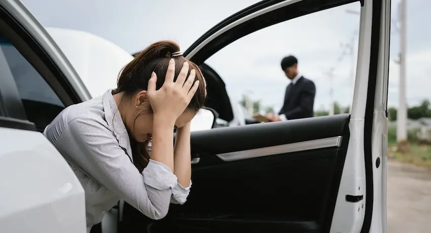 A distressed woman in a striped blazer sits in the open door of a white car with her head buried in her hands while a man in a dark suit writes on a clipboard in the background capturing the overwhelming stress accident victims feel when an old injury is aggravated in a new crash in Metro Atlanta.