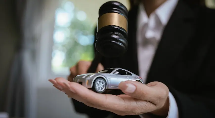 A lawyer from Hodgins & Kiber LLC in a black suit holds a silver toy car in an open palm while a wooden judge's gavel hovers directly above it symbolizing the legal proceedings involved when a car accident reopens or worsens an old injury and a victim seeks compensation in court.