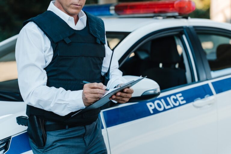 A police officer in a tactical vest writes notes on a clipboard while standing in front of a patrol car with flashing lights representing an officer documenting the details of an incident to complete an official police report in Atlanta.