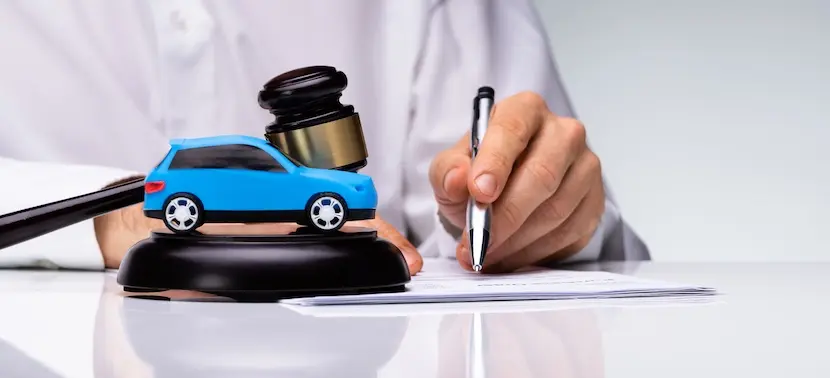 A person in a white shirt signs legal documents beside a judge's gavel resting on a small blue model car representing the process of filing a legal claim at Hodgins & Kiber LLC supported by an official police report following a vehicle accident in Atlanta.