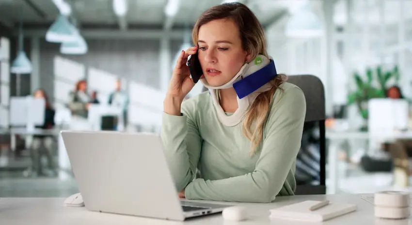 A blonde woman wearing a blue and white cervical neck brace sits at a desk in an open office talking on her phone with Hodgins & Kiber LLC while looking at a laptop representing an accident victim managing medical and legal matters at work after suffering a new trauma on top of an old injury in Metro Atlanta.