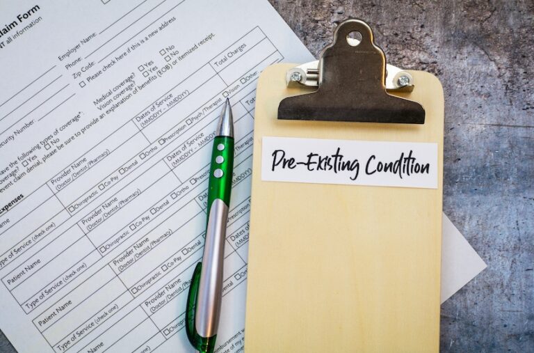 A wooden clipboard with a white label reading "Pre-Existing Condition" in handwritten script rests on top of a medical insurance claim form alongside a green and silver pen on a textured gray surface illustrating the documentation required when an accident aggravates an old injury and insurers dispute liability based on prior medical history in Metro Atlanta.