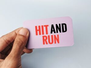 A hand holds up a small card printed with the words "Hit-and-Run" in bold red and black lettering directly referencing the criminal act and legal term associated with a hit-and-run accident in which a driver flees the scene in Atlanta.