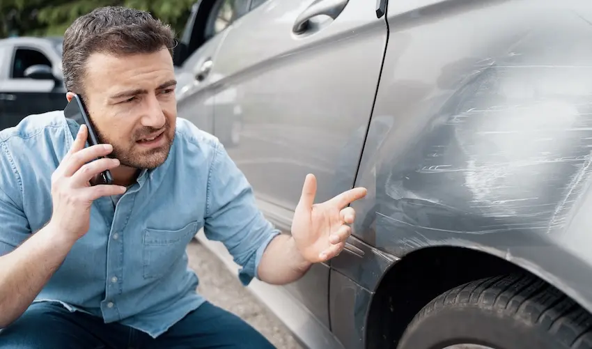 A frustrated man crouches beside his badly scraped and dented silver car while talking on his phone capturing the distress and urgency of a driver reporting the damage caused by a hit-and-run accident to Hodgins & Kiber LLC in Atlanta.