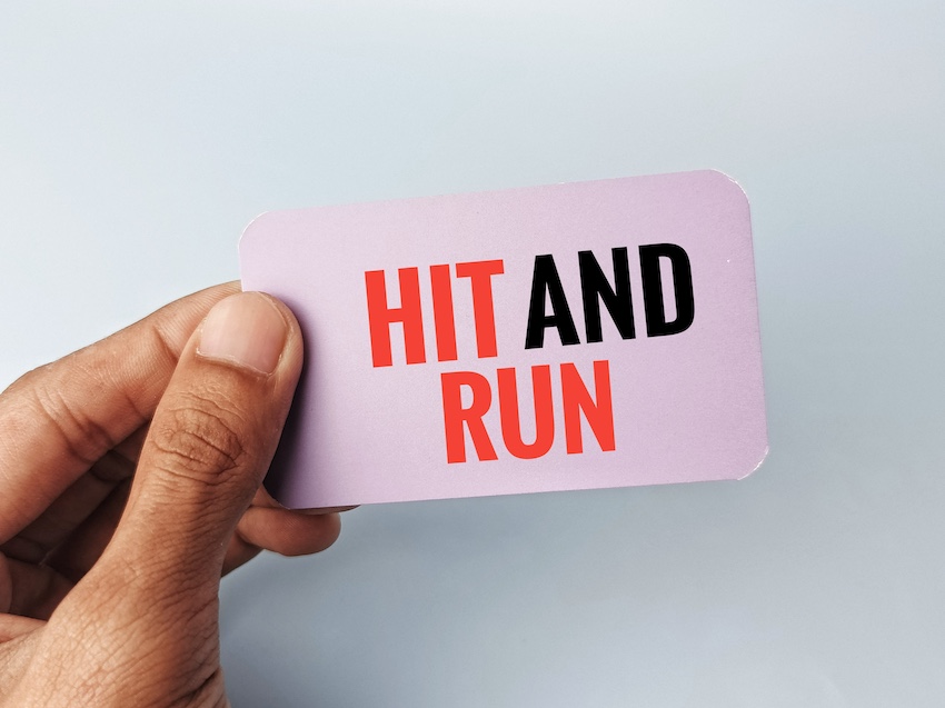 A hand holds up a small card printed with the words "Hit-and-Run" in bold red and black lettering directly referencing the criminal act and legal term associated with a hit-and-run accident in which a driver flees the scene in Atlanta.