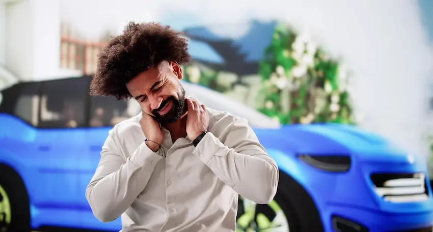 A man in a white dress shirt winces and grips both sides of his neck in pain while standing in front of a blue SUV illustrating the immediate neck and soft tissue discomfort that accident victims experience after being rear-ended by another vehicle.