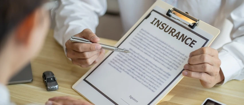 An attorney from Hodgins & Kiber LLC holds up a clipboard displaying an insurance document and points to specific terms with a pen as a client sits across the table beside a small toy car- showing the policy review process that determines how an insurance adjuster evaluates and settles a vehicle-related injury claim.