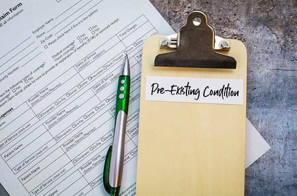 A wooden clipboard with a white label reading "Pre-Existing Condition" in handwritten script rests on top of a medical insurance claim form alongside a green and silver pen on a textured gray surface illustrating the documentation required when an accident aggravates an old injury and insurers dispute liability based on prior medical history in Metro Atlanta.