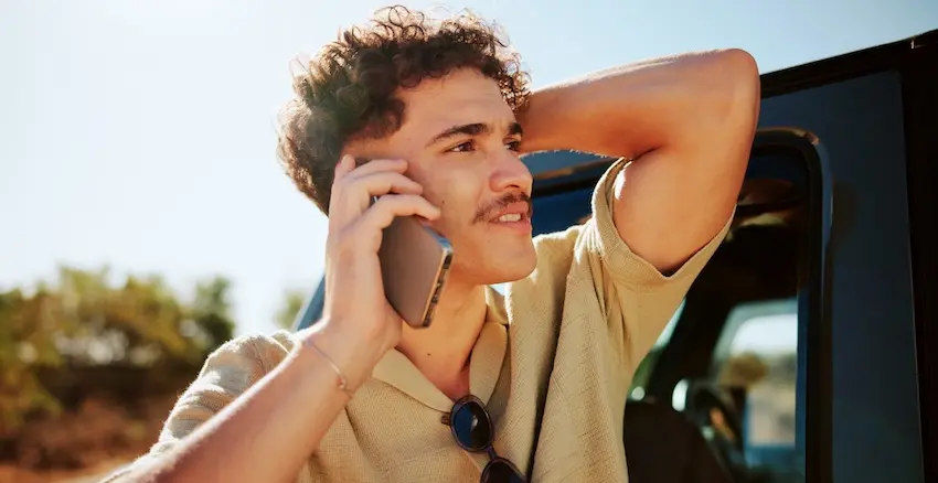 A young man with curly hair and a mustache in a beige linen shirt leans against a dark vehicle with sunglasses hanging from his collar talking on the phone with Hodgins & Kiber LLC and looking off into the distance on a sunny day- depicting one of the most important steps to take after a car accident which is calling an attorney to understand your legal rights before speaking with insurance.
