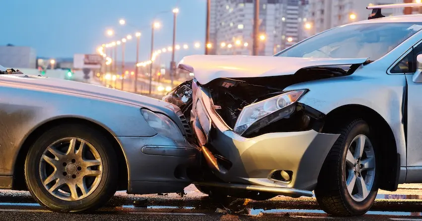 Two silver sedans with severely crumpled hoods locked together after a violent collision on a wet city road at dusk with glowing streetlights and a blurred urban skyline reflected in the pavement behind them illustrating the kind of serious property damage and potential injuries that require immediate legal and medical attention after a car accident in Metro Atlanta.
