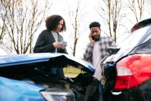 A woman with curly hair in a gray cardigan looks down at her phone while a man in a plaid shirt talks on his cell phone beside her- both standing between a blue car with a crumpled trunk and a black car with rear-end damage on a bare tree-lined road depicting two drivers exchanging information and contacting Hodgins & Kiber LLC after a car accident in Metro Atlanta.