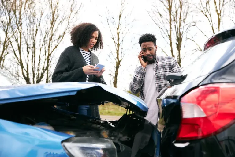 A woman with curly hair in a gray cardigan looks down at her phone while a man in a plaid shirt talks on his cell phone beside her- both standing between a blue car with a crumpled trunk and a black car with rear-end damage on a bare tree-lined road depicting two drivers exchanging information and contacting Hodgins & Kiber LLC after a car accident in Metro Atlanta.
