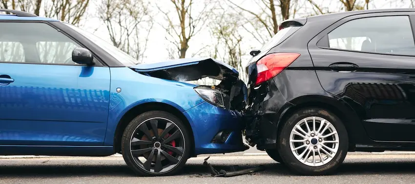 A bright blue hatchback with red brake calipers visible behind the front wheel has its entire hood crumpled upward and the front bumper torn away where it has rear-ended a black hatchback whose back end is crushed inward with the trunk lid buckled and deformed both vehicles locked together on a wet two-lane road with small pieces of plastic debris scattered on the pavement below the point of impact and a row of bare winter trees with pale gray branches stretching across the blurred background — the type of rear-end collision that requires victims to immediately contact their insurance company to begin the claims process and could use help from Hodgins & Kiber LLC.