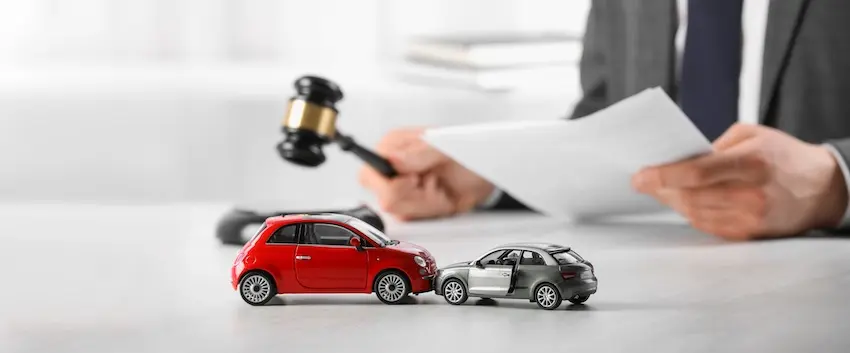 A lawyer in a gray business suit- from Hodgins & Kiber LLC- and dark blue tie sits at a white desk in the blurred background holding a black and gold judge's gavel in one hand and reviewing a white document in the other while in the sharp foreground a small red compact toy car and a silver SUV toy car are staged nose-to-rear in a collision with the silver vehicle's driver door popped open, positioned on the desk surface in front of the attorney — a staged legal concept image representing the role an attorney plays in negotiating with an insurance company on behalf of accident victims in Metro Atlanta.