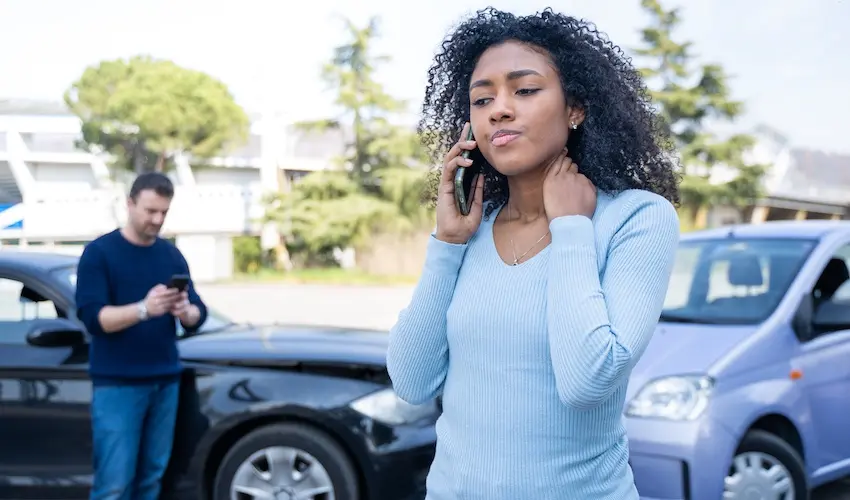 A young Black woman with voluminous natural curly hair wearing a light blue ribbed long-sleeve sweater stands in the foreground pressing a smartphone to her ear while reaching her other hand up to gingerly touch and hold the side of her neck- her expression tense and pained- suggesting a whiplash or neck injury sustained in the collision visible behind her where a black sedan with rear-end damage is parked and a man in a dark blue sweater stands in the background looking down at his own phone — capturing the immediate aftermath of a crash as the injured victim calls her insurance company to report the accident and seek guidance in Metro Atlanta.